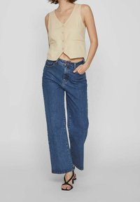 Béžová bezrukávová krátká vesta s knoflíky, kombinovaná se širokými modrými džínami. Oblečení má pohodlný střih a jednoduchý design.