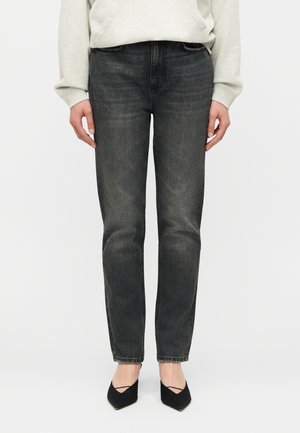 Moteris dėvi išblukusius juodus tiesių kojų džinsus, šviesiai pilką džemperį ir juodus aukštakulnius su smailiais pirštais, stovintį priešais baltą foną.