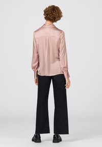 Ljusrosa satinblus med en söm bak, långa ärmar och krage, kombinerad med mörka vida jeans och svarta skor.