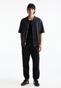 Zwarte kortekleurige button-up shirt in combinatie met zwarte cargo broek. De outfit heeft een relaxte pasvorm en een subtiele glans, aangevuld met zwarte sneakers.