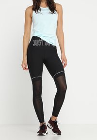 Vrouw in een lichtblauw mouwloos topje en zwarte leggings met de tekst "JUST DO IT", gecombineerd met maroon en witte sportschoenen.