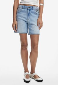Ljust blå denimshorts med normal passform, med blommönsterbroderi på framfickorna och en rak fåll. Kombinerat med vita slip-on-skor.