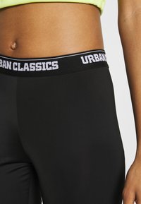 Juodos leggingai su plačiu elastingu juosmeniu, kuriame baltais įrašais parašyta "URBAN CLASSICS". Lygi tekstūra, prigludusi forma, vidutinio aukščio kirpimas.