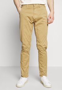 Bež chinos hlače iz bombaža, v slim fit kroju, z stranskimi žepe in gumbom za zapiranje v pasu. Ujemajo se z belimi supergami.