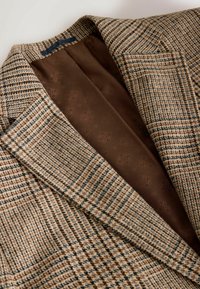 Brūns hounds-tooth žakets ar faktūru audumu, kas piedāvā gludu brūnu oderi ar smalkiem ziedu rakstiem. Ar izgrieztiem ragiem un pieguļošu piegriezumu.
