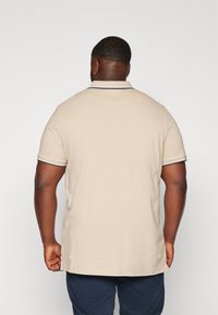 Beżowa koszulka polo z krótkimi rękawami, z kontrastującą czarną lamówką na kołnierzyku i prostym dołem. Materiał wydaje się miękki i lekko teksturowany.