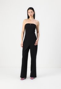 Sorte stroppeløse jumpsuit med glidelås foran, skreddersydd passform, utsvingte ben og glatt tekstur. Kombinert med lilla sandaler.