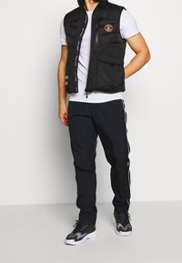 Man som bär svart vadderad väst, vit kortärmad skjorta, svarta träningsbyxor med vita ränder och svarta och vita sneakers, stående mot en vit vägg.