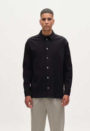 Black button-up shirt in textured fabric, featuring a collar and long sleeves, paired with light gray trousers. Buttons are visible and centered.