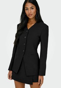 Zwart pak met een lange blazer met een V-hals en vijf knopen, gecombineerd met een korte rok. Glad materiaal en gestructureerd ontwerp.