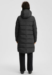 Persona, kas tērpta garā melnā pūkainā mētelī ar kapuci, melnās biksēs un melnās kurpēs, stāv ar muguru pret vienkrāsainu fonu.