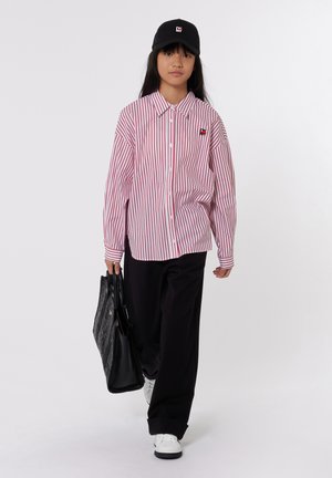 Chemise à rayures rouges et blanches, pantalon noir, baskets blanches et casquette noire. Porte un sac fourre-tout noir texturé.