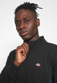 Black half-zip pullover made of smooth fabric, featuring a small branded logo patch on the chest and fine stitching details.