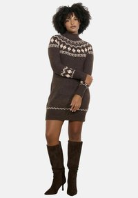 Abito maglione lavorato a maglia di colore marrone scuro con colletto alto, caratterizzato da motivi geometrici crema sulla scollatura e sulle maniche. Abbinato a stivali marroni alti fino al ginocchio.