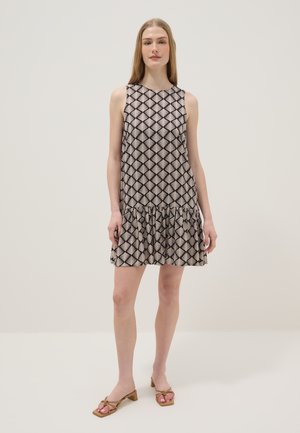 Frau steht und trägt ein ärmelloses, gemustertes Kleid mit Rüschensaum und beige flache Sandalen vor einem einfachen hellen Hintergrund.