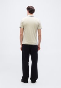 Un homme debout, de dos, portant un polo beige à manches courtes et un pantalon noir, sur un fond blanc uni.