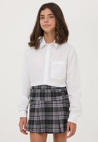 Camicia bianca con bottoni, a maniche lunghe e taschino sul petto, abbinata a una gonna a quadri nera con accenti viola e un dettaglio con fibbia laterale.