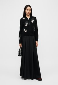 Crni kardigan na patentni zatvarač s apstraktnim vezenim licima; uparen s dugom crnom suknjom. Model drži malu crnu torbicu.