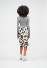 Femeie cu păr creț, îmbrăcată cu un top gri cu mâneci lungi, fustă până la genunchi cu model maro și alb, șosete albe și pantofi de culoare deschisă, stând în picioare.