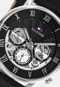 Orologio analogico nero e argento con design scheletrico, indicatori delle ore in numeri romani, display della data e del giorno, e un quadrante texture. Marchio: Hilfiger.
