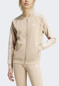 Chaqueta deportiva beige con cremallera y franjas blancas en las mangas; tela suave y lisa con puños acanalados. Combinada con leggings beige a juego.