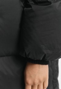 Zwarte pufferjack met een geborduurd patroon, voorzien van een comfortabele geribbelde manchette en subtiele branding op de mouw. Glad, lichtgewicht materiaal.