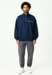 Polaire marine à demi-zip avec "Amour" brodé en blanc, portée avec un pantalon cargo gris et des baskets blanches. Coupe décontractée, poignets côtelés.