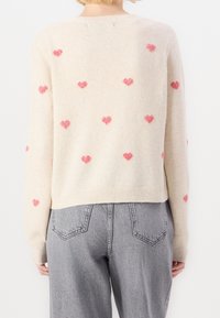 Beige stickad tröja med rosa hjärtmönster; rundad halsringning, ribbade kantar och manschetter. Bärs med grå högmidjade jeans. Texturen verkar mjuk.