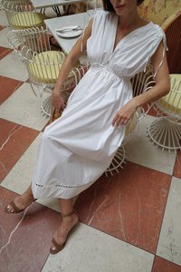 Robe en coton blanc avec des épaules froncées et une taille élastique. Finition en broderie au niveau de l'ourlet. Modèle assis sur un sol à motifs, portant des sandales beige.