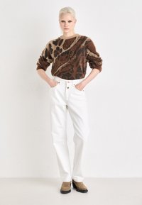 Suéter fuzzy de color marrón y negro con estampado, jeans blancos de talle alto y botas de tobillo marrones. La modelo posa con las manos en los bolsillos.
