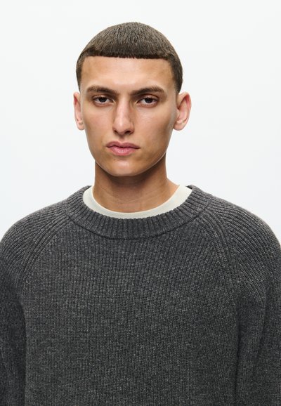 Ciemnoszara, dziana sweter o prążkowanej fakturze, z okrągłym dekoltem i rękawami raglanowymi. Pod spodem znajduje się jasna koszula.