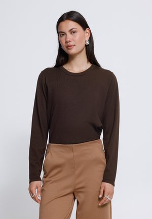 Femme aux longs cheveux foncés portant une chemise à manches longues marron foncé et un pantalon marron clair, debout avec une expression neutre sur un fond uni.