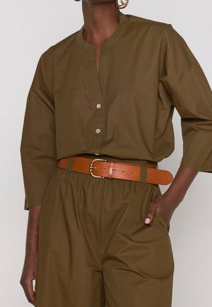 Kobieta ubrana w oliwkowozieloną koszulę na guziki i spodnie z szerokim jasnobrązowym skórzanym paskiem oraz złotymi kolczykami kołami, mająca rękę w kieszeni.