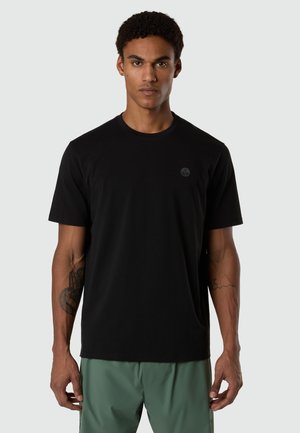 Uomo con capelli corti e ricci, che indossa una semplice maglietta nera a girocollo e pantaloni verdi, in piedi davanti a uno sfondo grigio.