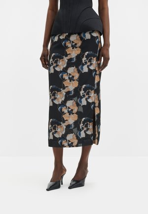 Femme portant une jupe midi noire avec de grands motifs floraux beige et bleu, associée à des mules noires à talons et un haut noir ajusté.
