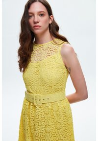 Gelbes Spitzenkleid mit floralem Muster, ärmellosem Design, hohem Ausschnitt und einem passenden Gürtel in der Taille. Glatte Textur.