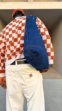 Jeansowa torba na ramię o zaokrąglonym kształcie, ciemnoniebieskim kolorze i kontrastujących przeszyciach. Noszona z wzorzystym swetrem i jasnymi spodniami.