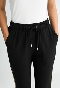 Czarne spodnie dresowe z elastycznym pasem i sznurkiem, kieszeniami po bokach oraz złotymi zdobieniami na nogawkach. Gładka faktura tkaniny.