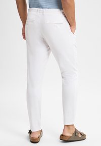 Witte slim-fit broek van een geweven stof. Bevat twee achterzakken met knoopsluitingen. Gemaakt om te dragen met bruine slippers.
