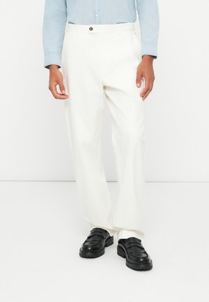 Weiße Baumwollhosen mit lockerer Passform, ausgestattet mit einem vorderen Knopfverschluss und Seitentaschen, kombiniert mit schwarzen Schuhen. Strukturierter Stoff.