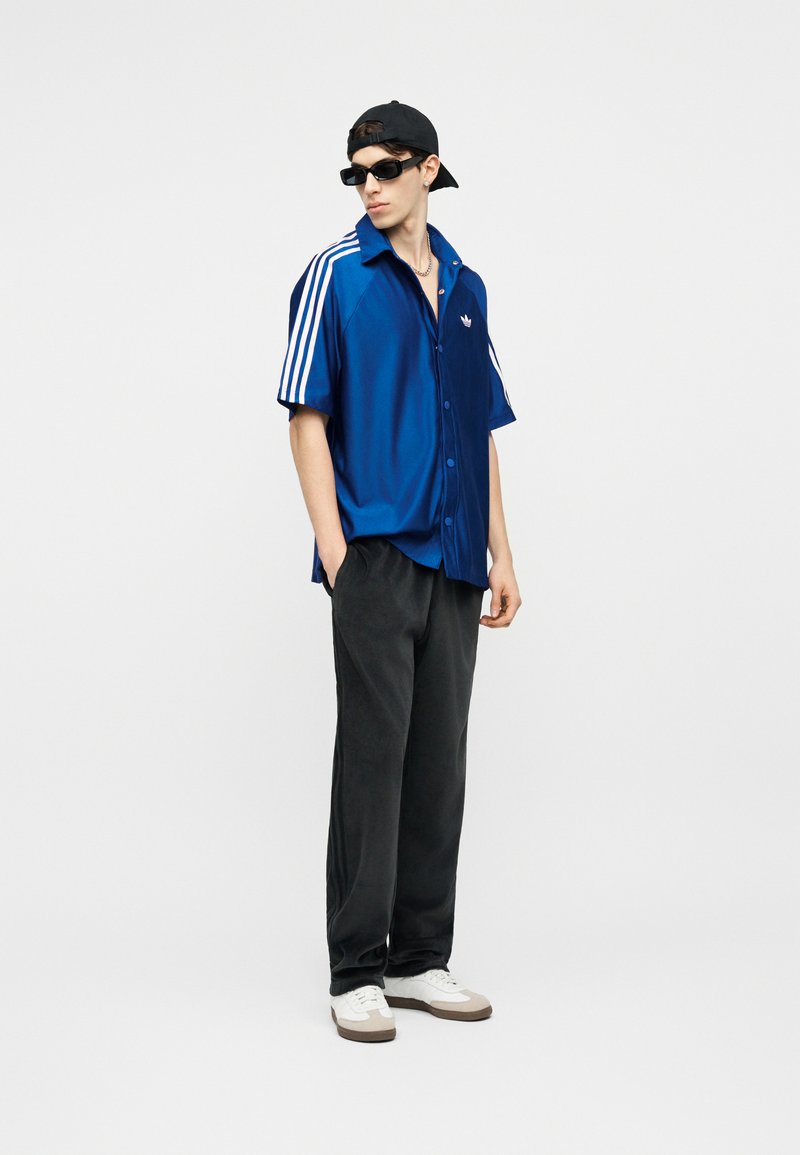 Giovane uomo che indossa una maglietta blu Adidas con strisce bianche, pantaloni neri, scarpe da ginnastica bianche, cappellino nero indossato al contrario e occhiali da sole neri, in piedi con le mani nelle tasche.