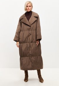 Cappotto lungo imbottito marrone con ampio colletto, silhouette oversize e vita elasticizzata, abbinato a stivali con tacco alto in stampa leopardata.