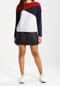 Sudadera con bloques de color en gris, azul marino y rojo con mangas largas, combinada con una mini falda de cuero negra con botones frontales. Zapatillas blancas.