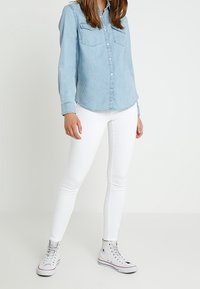 Camicia di denim azzurro chiaro con maniche lunghe e tasche frontali, abbinata a jeans skinny bianchi e sneakers grigie alte.