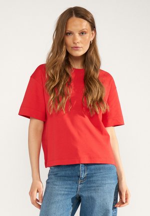 Vrouw met lang golvend bruin haar draagt een losse rode korte mouwen t-shirt en blauwe spijkerbroek, staand tegen een egale lichte achtergrond.
