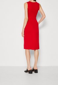 Robe midi rouge moulante sans manches avec texture côtelée et fente au dos ; associée à des mules noires à talon bloc.