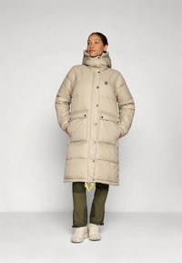 Beige padded long coat with a high collar, button closure, and two front pockets. Paired with olive green pants and white sneakers.