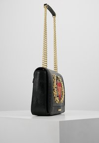 Svart läderaxelväska med guldkedjehandtag, prydlig med röd och guld dekorativ pärlbroderi på frontpanelen, utställd på en vit plattform.