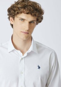 Giovane uomo con capelli ricci castani che indossa una camicia bianca con bottoni e un piccolo logo ricamato blu scuro sul petto.