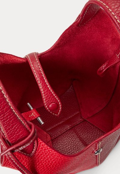 Interiér červenej koženej kabelky zobrazujúci štruktúru zrna, prešitý popruh s gombíkovým zapínaním a hladké červené semišové obloženie.